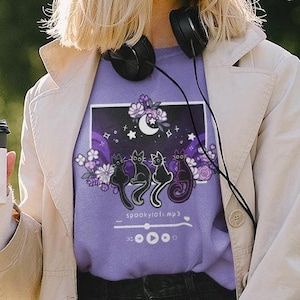 May include: A woman wearing a light purple t-shirt with a graphic of three black cats, a crescent moon, and stars. The graphic is surrounded by purple flowers and the text "spookyvibes.mp3".