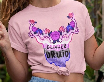 Könnte beinhalten: Ein pinkfarbenes T-Shirt mit einem Aufdruck eines Hirschkopfes mit Rosenranken als Geweih. Der Hirschkopf ist in den Farben der bisexuellen Pride-Flagge gehalten. Unter dem Hirschkopf steht der Text "Gender Druid".