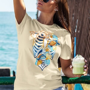 May include: A woman wearing a light yellow t-shirt with a graphic of a ribcage filled with seashells and a turtle. She is wearing blue denim shorts and sunglasses. She is holding a smoothie in her right hand.