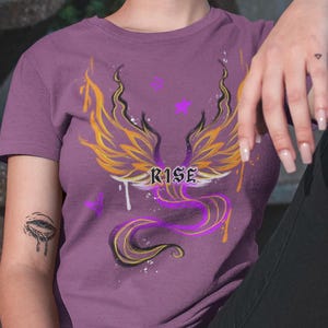 May include: A purple t-shirt with a graphic of a phoenix with yellow and purple wings and the word "RISE" in the center. The phoenix is surrounded by purple and yellow flames and stars.