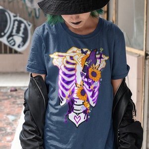 Peut inclure: Un t-shirt bleu marine avec un motif de squelette avec des tournesols et un cœur violet. Le squelette porte une couronne et a une chauve-souris sur la tête.