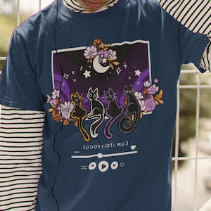 Peut inclure: Un t-shirt bleu marine avec un motif de trois chats noirs assis devant un croissant de lune et des étoiles. Le motif comprend également des fleurs et le texte "spookylofi.mp3" avec une icône de lecteur de musique.