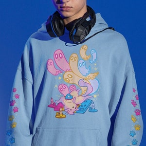 Peut inclure: Un sweat-shirt bleu clair avec un imprim&eacute; graphique d'un chat portant un casque et des fant&ocirc;mes. Les manches ont un motif d'empreintes de pattes en bleu, jaune et rose.