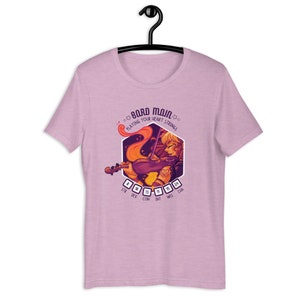 May include: A pink t-shirt with a graphic of a woman playing a violin. The graphic is surrounded by a hexagon with the text "Bard Main" and "Playing Your Heart Strings". The hexagon also has a set of dice with numbers representing different stats: STR, DEX, CON, INT, WIS, CHA.