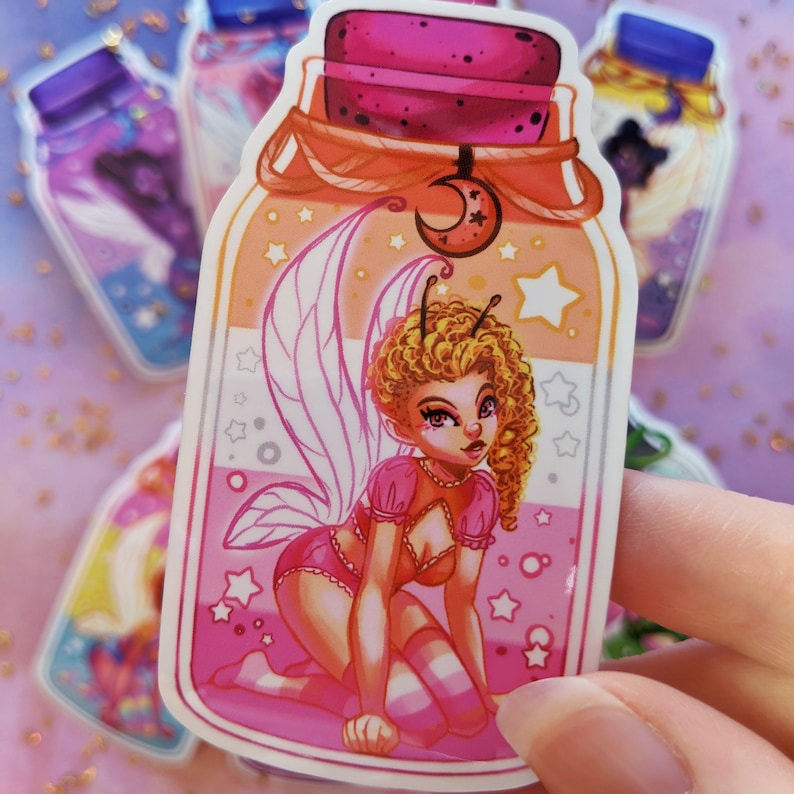 May include: A sticker of a fairy with pink wings and a pink dress inside a glass jar with a pink, orange, and white background. The jar has a cork lid and a crescent moon charm.