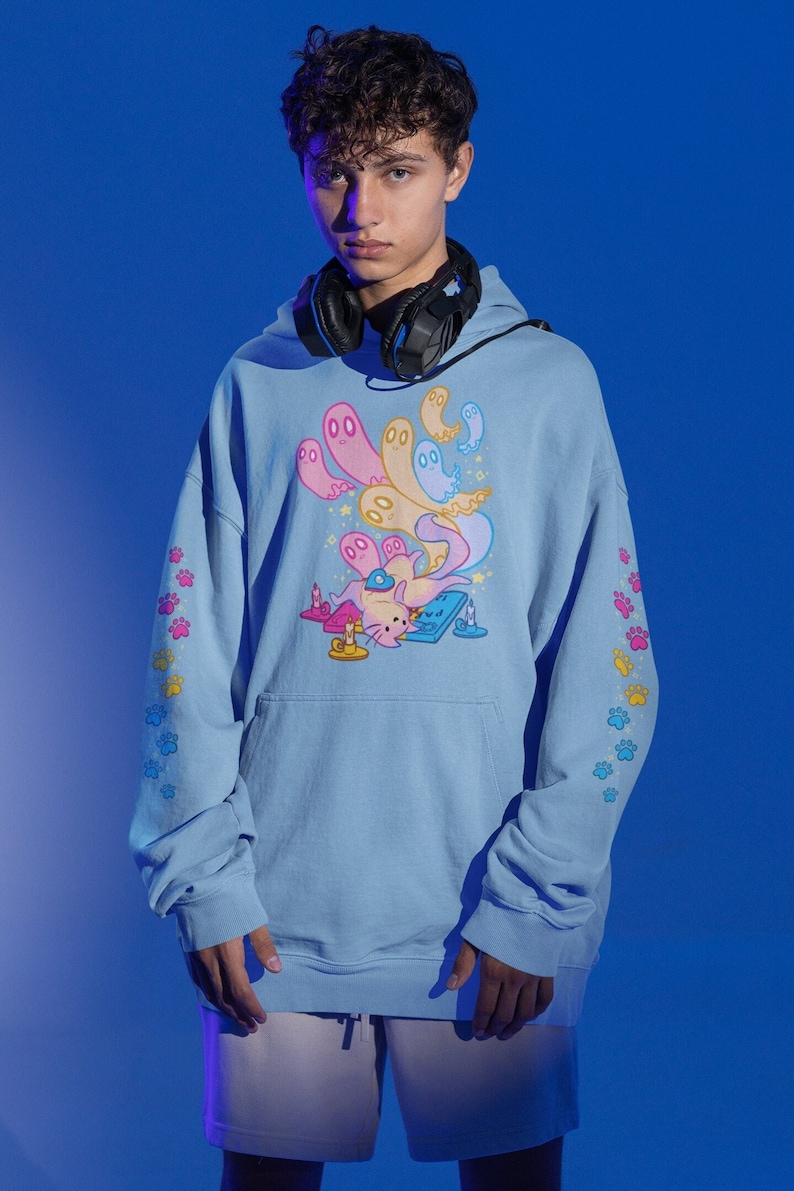 Peut inclure: Un sweat-shirt bleu clair avec un imprim&eacute; graphique d'un chat portant un casque et des fant&ocirc;mes. Les manches ont un motif d'empreintes de pattes en bleu, jaune et rose.