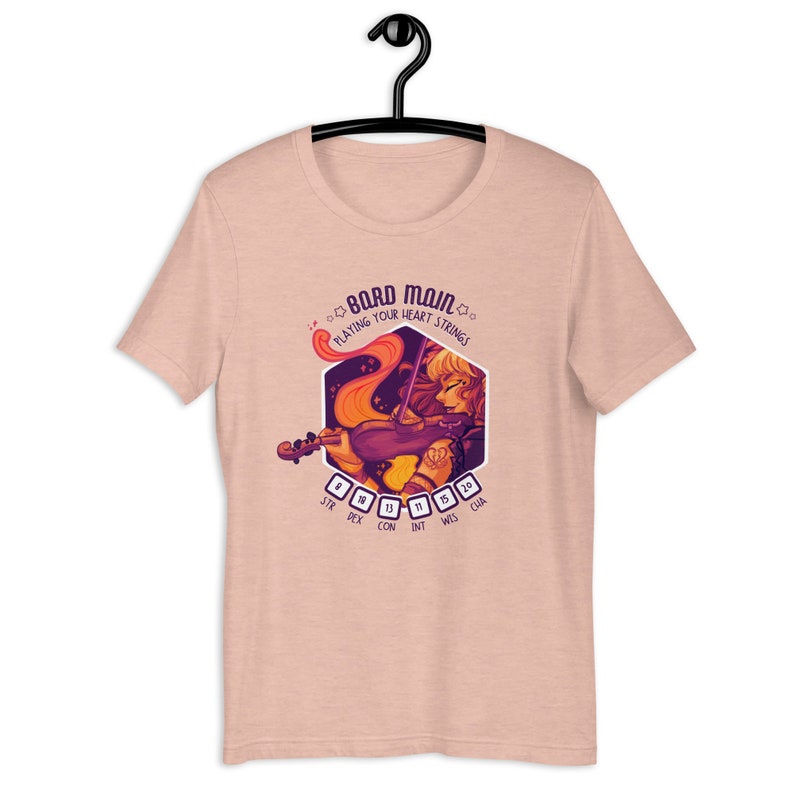 May include: A pink t-shirt with a graphic of a woman playing a violin. The graphic is in a hexagon shape with the text "Bard Main" and "Playing Your Heart Strings" above the image. The text "STR DEX CON INT WIS CHA" is below the image with dice numbers next to each word.
