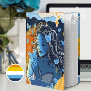 Puede incluir: Un cuaderno de tapa dura con una ilustración gráfica de una figura de piel azul con cabello largo y oscuro rodeada de mariposas azul claro. El diseño de la portada incluye detalles en naranja y amarillo, formas de estrellas y un patrón de cuadrícula.