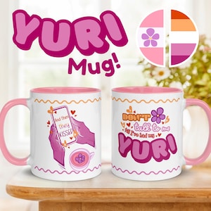 May include: Two white ceramic mugs with pink handles and interiors. One mug features the text "And then they kissed" with a phone graphic. The other mug says "Don't talk to me till I've had my Yuri." Both mugs have a pink and orange wavy design.