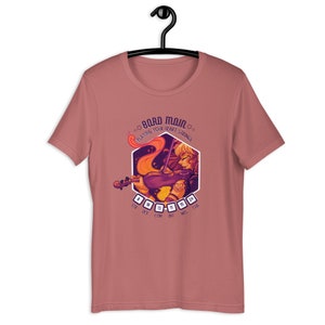 May include: A pink t-shirt with a graphic of a woman playing a violin. The graphic is surrounded by a hexagon with the text "Bard Main" and "Playing Your Heart Strings". The hexagon also has dice with numbers representing stats: STR, DEX, CON, INT, WIS, CHA.