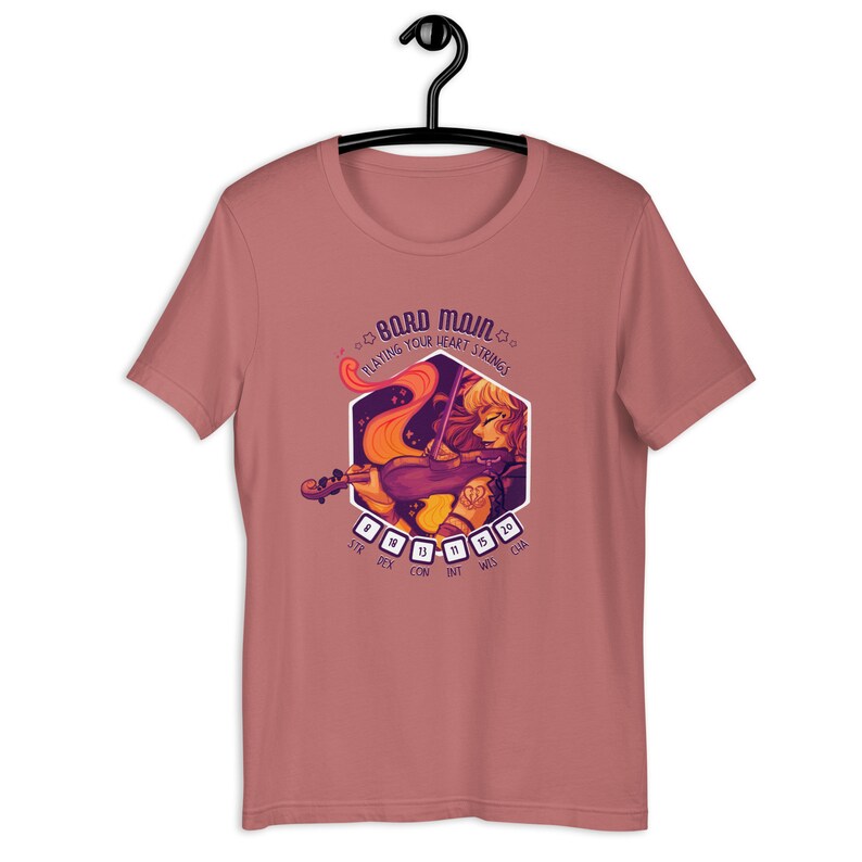 May include: A pink t-shirt with a graphic of a woman playing a violin. The graphic is surrounded by a hexagon with the text "Bard Main" and "Playing Your Heart Strings". The hexagon also has dice with numbers representing stats: STR, DEX, CON, INT, WIS, CHA.