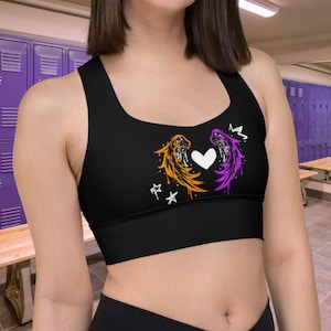 May include: Black sports bra featuring a design with orange and purple wings, a white heart, and stars. The bra has a scoop neckline and wide straps. The design is centered on the front of the bra.