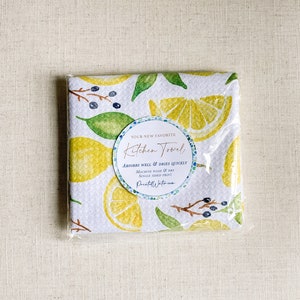 May include: A white kitchen towel with a lemon and leaf pattern. The towel has a label that says "Your New Favorite Kitchen Towel" and "Absorbs Well & Dries Quickly".