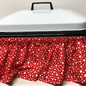 May include: A white appliance with a black handle and trim, featuring a red skirt with a white star pattern. The skirt is gathered and ruffled, adding a decorative element to the appliance.