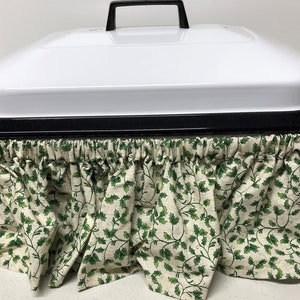 May include: A white appliance with a black handle and trim, featuring a ruffled skirt. The skirt is made of fabric with a cream background and a green holly leaf pattern. The appliance is likely for kitchen use.