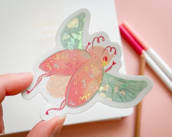 May include: A pink and green iridescent beetle sticker with gold glitter accents. The beetle has a whimsical design with a large body and small legs.