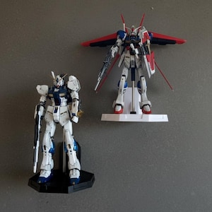 May include: Two detailed Gundam model kits. One is white, blue, and gold, standing on a black base. The other is white, blue, and red, with large wings and a shield, mounted on a white stand. Both are posed against a gray background.
