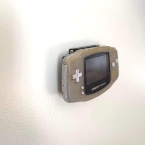May include: A Game Boy Advance handheld console, mounted on a white wall. The console is a light gray color with a black screen and a directional pad. The text "GAME BOY ADVANCE" is visible.