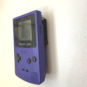 May include: A purple Game Boy Color handheld game console mounted on a wall. The console features a screen, directional pad, buttons, and the text "GAME BOY COLOR" in white. The console is attached to a black bracket.