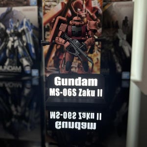 Pode incluir: Um kit de modelo Gundam MS-06S Zaku II vermelho e marrom, segurando um rifle, está em frente a uma tela com o texto "Gundam MS-06S Zaku II". O modelo é cercado por outras caixas de modelos Gundam.