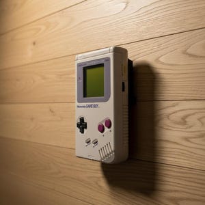 May include: A vintage Nintendo Game Boy handheld console mounted on a light wood-paneled wall. The Game Boy is light gray with a green screen, purple accents, and the text "Nintendo GAME BOY." The console's buttons are black and purple.