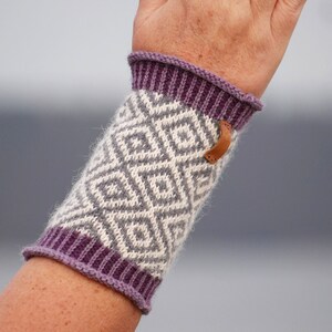 Può includere: Scaldamani in maglia viola e bianca con un motivo geometrico. Gli scaldamani hanno un bottone in pelle marrone.