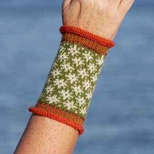 May include: A green and white knitted wrist warmer with an orange trim. The wrist warmer is on a person's arm, and the person is wearing a silver ring with a white stone.