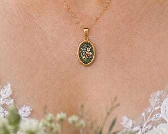 May include: A gold chain necklace featuring an oval pendant with a dark green background and a small, multi-colored floral embroidery design. The pendant is framed in a polished gold-tone metal setting, suspended from a delicate gold link chain.