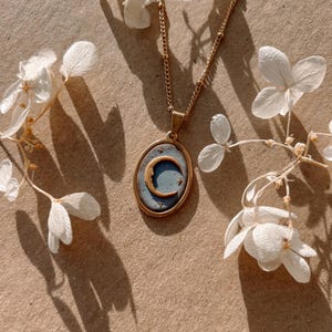 May include: A gold-toned necklace featuring an oval pendant with a matte blue center. The pendant displays a raised gold crescent moon and small star accents. The necklace is suspended from a delicate gold-toned chain with small bead details.