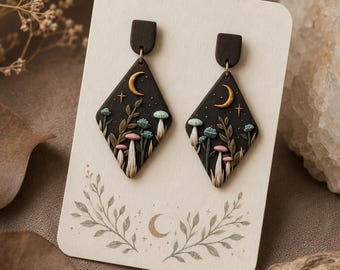 May include: A pair of diamond-shaped earrings featuring a black base with raised, colorful mushroom and foliage designs. Each earring displays a gold crescent moon and small stars. The earrings are attached to a cream-colored display card.