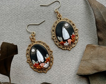May include: A pair of gold-toned earrings with a black background and a white ghost figure. The ghost is standing in front of a small orange and white flower arrangement. The earrings are oval shaped and have a decorative gold frame.