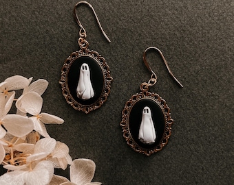 Puede incluir: Un par de pendientes de tono dorado con colgantes de camafeo de fantasma blanco y negro. El fantasma es blanco y tiene un contorno negro. Los pendientes tienen forma ovalada y tienen un borde decorativo.
