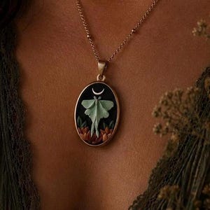 May include: An oval pendant necklace featuring a pale green moth centered on a black background, topped with a small silver crescent moon. The base of the pendant displays orange and brown floral elements, all encased in a gold-toned metal frame.