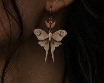 Può includere: Un orecchino pendente raffigurante una falena color crema con intricati motivi sulle ali, sospeso a un piccolo ciondolo a forma di mezzaluna color oro. La falena presenta un corpo centrale e lunghe ali inferiori appuntite.
