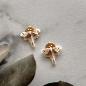 May include: A pair of white and gold earrings featuring a delicate moth design. The moths have intricate wings and a gold accent on the body.