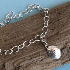 Könnte beinhalten: Ein silbernes Kettenarmband mit einem silbernen Muschel-Anhänger. Das Armband liegt auf einem Stück Treibholz.
