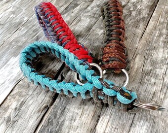 Puede incluir: Tres llaveros fabricados con paracord trenzado en distintas combinaciones de colores. Uno presenta rojo brillante y azul, otro es de color marrón oscuro sólido y el tercero es una mezcla de turquesa claro y gris oscuro, cada uno unido a una anilla de metal.