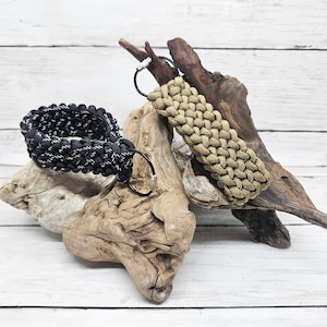 May include: Two paracord keychains. One is black with white accents, the other is tan. Both are braided and feature a metal keyring. The keychains are displayed on driftwood against a white wood background.