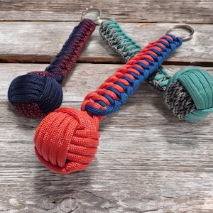 May include: A collection of handmade paracord keychains in various colors. The keychains feature a knotted ball design with braided handles. Colors include red, blue, teal, and a mix of black and gray. Each keychain has a silver metal ring.