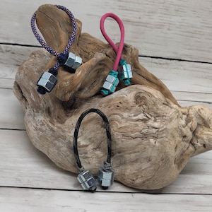 May include: Three zipper pulls made of looped cord and silver-toned metal hex nuts. One pull features a purple and black patterned cord, another has a solid pink cord with teal accents, and the third uses a black cord with white flecks.