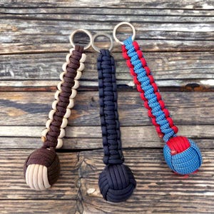 May include: Three handmade keychains with metal rings. One is brown and tan, one is navy blue, and the third is red and blue. Each keychain features a knotted design and a round, decorative end.
