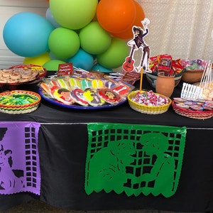 May include: A table decorated for a party with a black tablecloth, colorful balloons, and various treats. There are paper cutouts of characters from the movie Coco, including Miguel, Hector, and Dante. The table is set with a variety of snacks, including cookies, candy, and fruit.