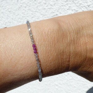 Puede incluir: Una delicada pulsera de cuentas con pequeñas cuentas grises facetadas y tres vibrantes cuentas rosas. La pulsera se lleva en la muñeca, mostrando el contraste de colores y texturas. Una pieza de joyería sencilla y elegante.