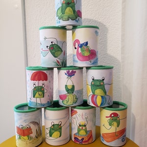 May include: A stack of 10 green and white cylindrical containers with a green rim. Each container features a different illustration of a frog engaging in various summer activities, such as reading a book, riding a surfboard, and holding an umbrella.