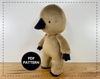 May include: A plush toy in the shape of a bird with a black beak and black eyes. The toy is brown and has a simple design. The toy is standing on a wooden surface. The text "PDF PATTERN" is on a black circle in the lower left corner of the image.