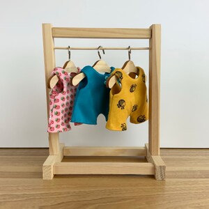 May include: A wooden doll clothes rack with three hangers holding doll clothes. The clothes are a pink and white strawberry print, a blue overall, and a yellow overall with a bee print.