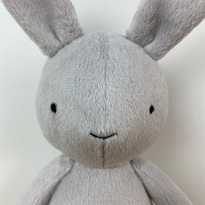 May include: A grey plush bunny with black embroidered eyes and a small smile. The bunny has long floppy ears.