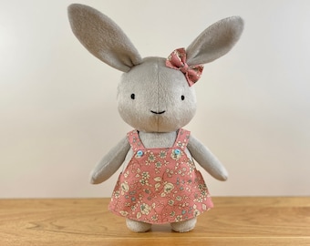 May include: A gray plush bunny wearing a pink floral dress with a pink bow. The bunny has black eyes and a button nose.