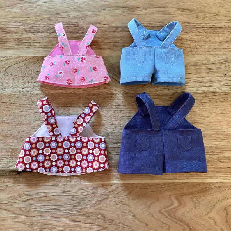 K&ouml;nnte beinhalten: Vier verschiedene Puppenoutfits, zwei mit Blumenmustern und zwei mit Jeans. Die Blumenoutfits sind pink und rot mit wei&szlig;en Akzenten. Die Jeansoutfits sind blau und dunkelblau.