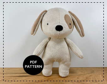 May include: A beige plush dog toy with brown ears and a black nose. The toy has a simple design with black eyes and a brown spot on one ear. The toy is standing on a wooden surface. The text "PDF PATTERN" is on a black circle in the lower left corner of the image.
